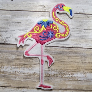 May include: Pink flamingo embroidered patch with yellow and blue floral details. The flamingo is standing on one leg with its head turned to the side.