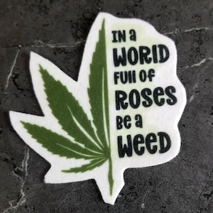 Peut inclure: Un patch en feutre blanc en forme de feuille de cannabis avec un motif de feuille de cannabis verte sur le côté gauche. Le côté droit porte le texte "IN A WORLD FULL OF ROSES BE A WEED" en noir.