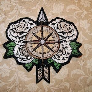 May include: Embroidered patch featuring a compass with a brown and gold design surrounded by white roses with green leaves. The compass has a red needle pointing north and a white center. The patch is on a beige background.