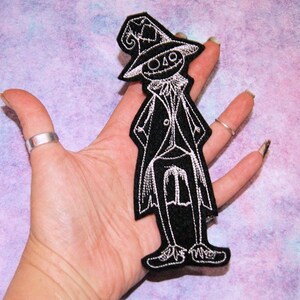 May include: Black and white embroidered iron-on patch of a spooky character wearing a tall hat and a long coat. The character has a stitched-on smile and a pointed hat.