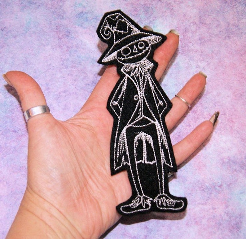 May include: Black and white embroidered iron-on patch of a spooky character wearing a tall hat and a long coat. The character has a stitched-on smile and a pointed hat.