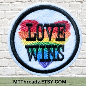 May include: A round, white patch with a black border and a rainbow heart. The heart has the words "LOVE WINS" embroidered in black.