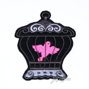 May include: Black embroidered iron cage with a pink bird inside. The cage has a decorative top with swirls and a base with a scalloped edge.