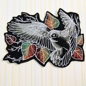 May include: Embroidered iron-on patch featuring a white bird with outstretched wings flying through a branch of autumn leaves. The leaves are a mix of red, orange, and green.