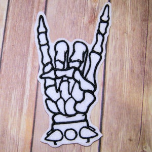 May include: A white embroidered patch of a skeleton hand making the rock and roll hand gesture. The patch is outlined in black thread.