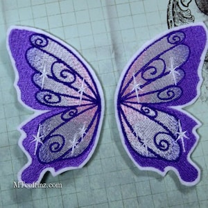 May include: A pair of purple and white embroidered fairy wings with a white border. The wings have a sparkly design and are perfect for adding a touch of magic to any costume or craft project.