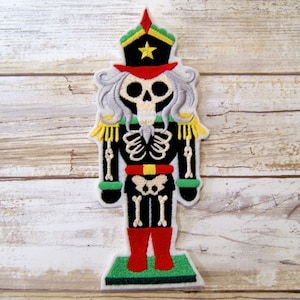 May include: A black and white embroidered patch of a skeleton nutcracker. The nutcracker is wearing a red and green hat with a yellow star. The nutcracker is holding a red and yellow stick in its right hand and a green stick in its left hand.