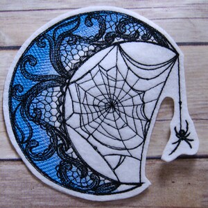 May include: Embroidered patch featuring a black and white spider web with a spider hanging from a strand of web. The web is set against a blue and white crescent moon design.