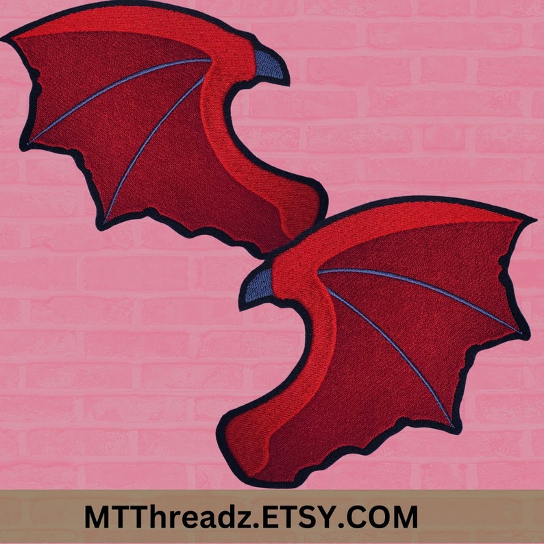 Puede incluir: Dos alas de tela bordadas de color rojo con contornos negros y costuras azules. Las alas est&aacute;n sobre un fondo de ladrillos rosas. MTTThreadz.ETSY.COM est&aacute; impreso en la parte inferior de la imagen.