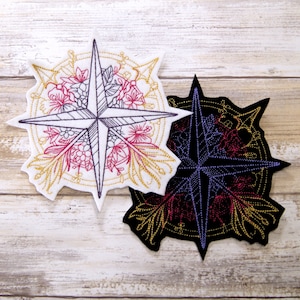 May include: Two embroidered patches featuring a compass rose design. The first patch is white with a gold outline and pink and red floral details. The second patch is black with a gold outline and red and purple floral details.