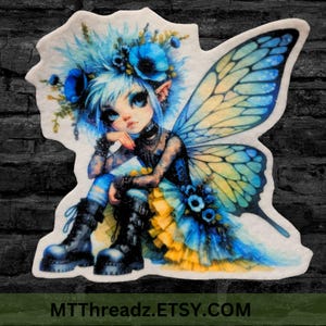 May include: A blue and yellow fairy with large wings and blue flowers in her hair. She is wearing black clothing and boots. The fairy is sitting on a white background. MTTThreadz.ETSY.COM is written at the bottom of the image.