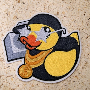 May include: Embroidered patch featuring a yellow duck wearing a black cap, star-shaped sunglasses, and a gold chain with a dollar sign pendant. The duck is holding a silver boombox. The patch has a white border.