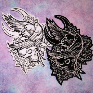 May include: Two embroidered patches featuring a black crow perched on a skull with floral accents. One patch is white with black embroidery, the other is black with white embroidery.