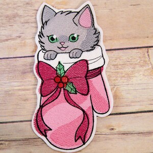 May include: Embroidered patch featuring a gray cat with green eyes peeking out of a pink mitten. The mitten has a large red bow with holly and berries. The patch has a white border.