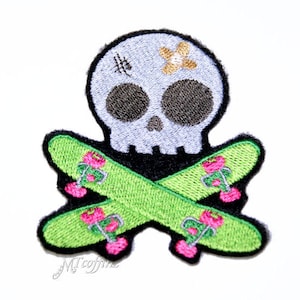 May include: A white skull with a black outline and a small yellow flower on its forehead is crossed with two green skateboards with pink wheels. The skateboards are embroidered with pink flowers.