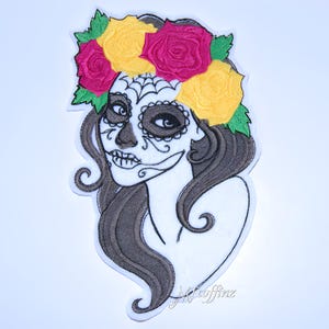 May include: Embroidered iron-on patch featuring a Day of the Dead sugar skull woman with a floral crown of yellow and red roses. The skull has a white face with black eyes and a black spiderweb design.