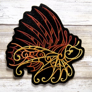 Peut inclure: Un patch brodé thermocollant représentant un poisson-lion stylisé avec des détails orange, rouge et jaune sur un fond noir.