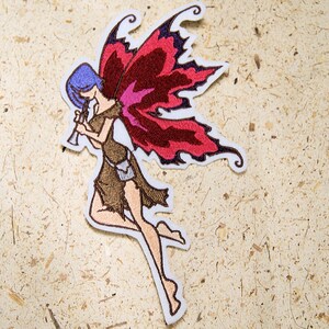 May include: Embroidered iron-on patch featuring a fairy with red and pink wings playing a flute. The fairy has brown hair and is wearing a brown outfit.