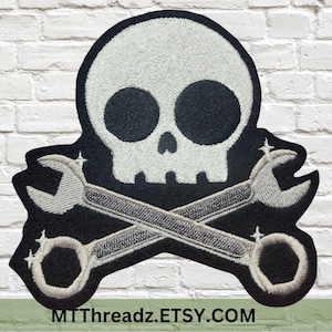 May include: Black and white embroidered patch featuring a skull and crossbones with two wrenches. The skull has large black eyes and a white outline. The wrenches are silver with a black outline and have a star-shaped detail on each side.