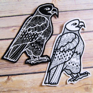 May include: Two embroidered eagle patches. One is black with white detailing, the other is white with black detailing. Both patches feature intricate patterns and are set against a wooden background.