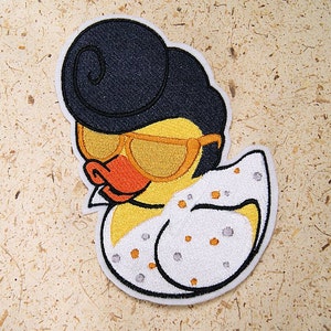 May include: Embroidered iron-on patch of a yellow rubber duck wearing sunglasses and a white jumpsuit with black polka dots. The duck has a black pompadour hairstyle.