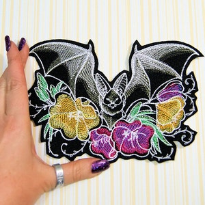 May include: Embroidered black patch with a bat design, detailed wings, and colourful flowers in yellow, purple, and green. The patch features intricate stitching and a white outline, creating a striking contrast.