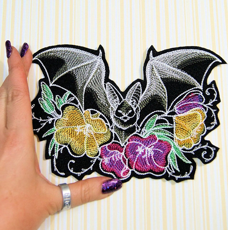 May include: Embroidered black patch with a bat design, detailed wings, and colourful flowers in yellow, purple, and green. The patch features intricate stitching and a white outline, creating a striking contrast.
