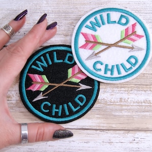 May include: Two embroidered patches with the text "WILD CHILD" in a white circle with a turquoise border and a black circle with a turquoise border. The patches feature two crossed arrows with pink, green, and gold accents.