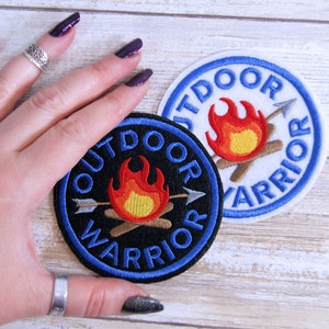 May include: Two embroidered patches with a blue border and the text "Outdoor Warrior" in white. The patches feature a campfire with an arrow through it. One patch has a black background and the other has a white background.