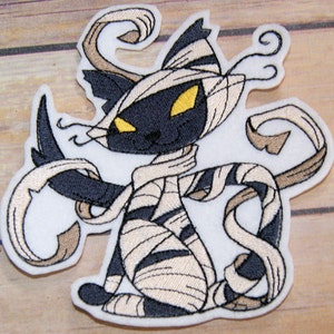May include: A black and white embroidered patch of a cat dressed as a mummy. The cat has yellow eyes and is wearing white bandages. The cat's tail is curled around its body.
