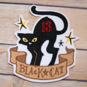 May include: Embroidered iron-on patch featuring a black cat with yellow eyes, a red number "13" on its back, and a brown banner that reads "BLACK CAT". Two yellow stars are in the background.