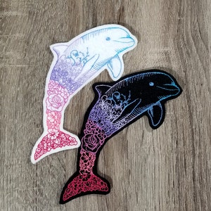 Peut inclure: Deux patchs thermocollants brodés de dauphins avec des motifs floraux. Le dauphin de gauche est blanc avec un motif floral rose et bleu. Le dauphin de droite est noir avec un motif floral violet et rose.