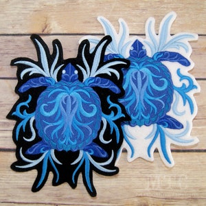 May include: Two embroidered iron-on patches featuring a blue and white stylized sea turtle with intricate designs. The patches are on a wooden background.