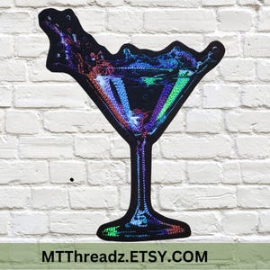 May include: A colorful, iridescent martini glass with a splash of liquid. The glass is made of sequins and has a black background.