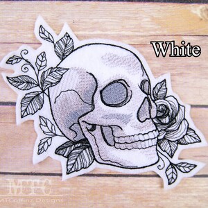 May include: White embroidered patch featuring a skull with a rose and leaves. The skull is outlined in black and has a detailed design. The rose is also outlined in black and has a detailed design. The leaves are also outlined in black and have a detailed design.