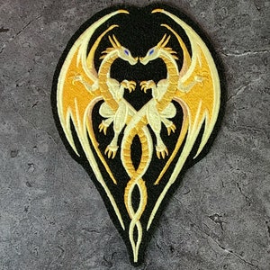 May include: A black embroidered patch featuring two gold dragons intertwined in a heart shape. The dragons have yellow wings and blue eyes.
