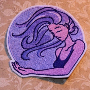 May include: Purple embroidered patch of a woman with long flowing hair, with her eyes closed, in a circle.