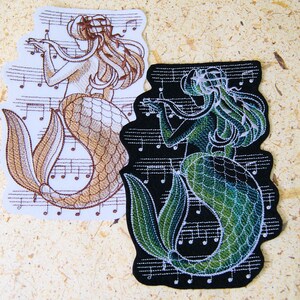 May include: Two embroidered patches of mermaids playing musical instruments. The mermaid on the left is brown and the mermaid on the right is green.