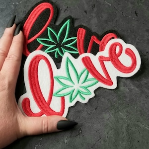 Puede incluir: Parche de fieltro rojo y blanco con la palabra "Love" bordada en rojo. El parche tiene dos diseños de hojas de marihuana verdes bordadas.