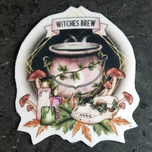 May include: A white patch with a hand-painted illustration of a cauldron with steaming liquid, surrounded by mushrooms, candles, leaves, and a skull. The text "WITCHES BREW" is written on a banner above the cauldron.