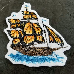 May include: A white fabric patch with an embroidered image of a ship made of monarch butterfly wings sailing on blue water.