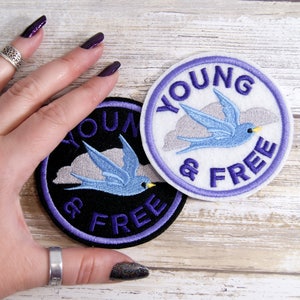 May include: Two round embroidered patches with the text "YOUNG & FREE". One patch is black with a purple border, the other is white with a purple border. Both patches feature a blue bird flying in front of a gray cloud.