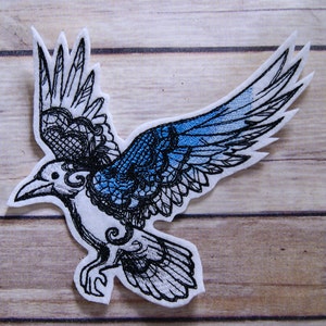 May include: A white embroidered patch of a black crow with blue wings. The crow is in flight with its wings spread wide. The crow's wings are detailed with a lace-like pattern.