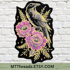 May include: A black fabric patch featuring an embroidered design of a raven perched above two vibrant pink flowers with gold outlines and foliage. Small gold stars are scattered around the bird and flowers. The text MTTTHREADZ.ETSY.COM is visible.