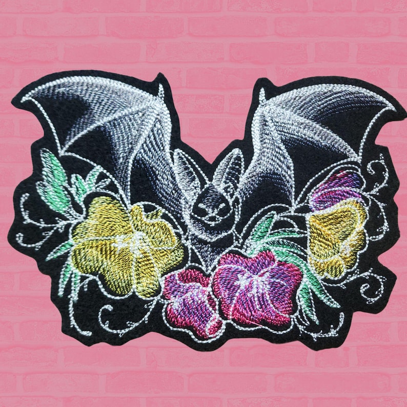 May include: Embroidered patch featuring a bat with outstretched wings, detailed with white stitching. The bat is surrounded by colourful flowers in yellow, purple, and pink, with green leaves. The patch is on a black background.