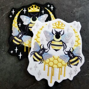 May include: Two embroidered patches featuring a bee with a crown, surrounded by a crescent moon and honeycomb dripping with honey. The patches are on a white and black background.