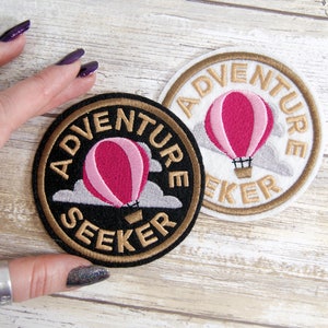 May include: Two embroidered iron-on patches with a hot air balloon design. The patches are round and have a gold outline. One patch is black with gold lettering and the other is white with gold lettering. Both patches say "Adventure Seeker".