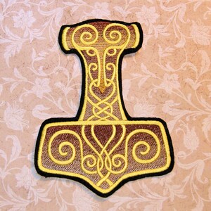 May include: A gold embroidered patch depicting Mjolnir, Thor's hammer, with intricate knotwork designs. The patch is on a brown fabric background.