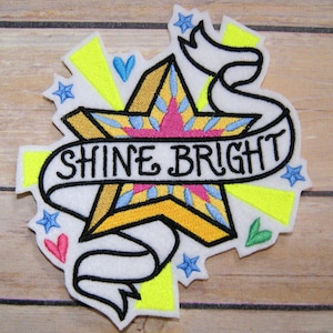 May include: A white felt patch with a yellow star and a white banner that says "Shine Bright". The star has a pink and blue design. The banner is surrounded by yellow and blue stars and hearts.