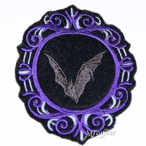 May include: Purple and white embroidered patch with a black background and a gray bat in the center. The bat is inside a circle with a purple border. The circle is surrounded by a decorative frame.